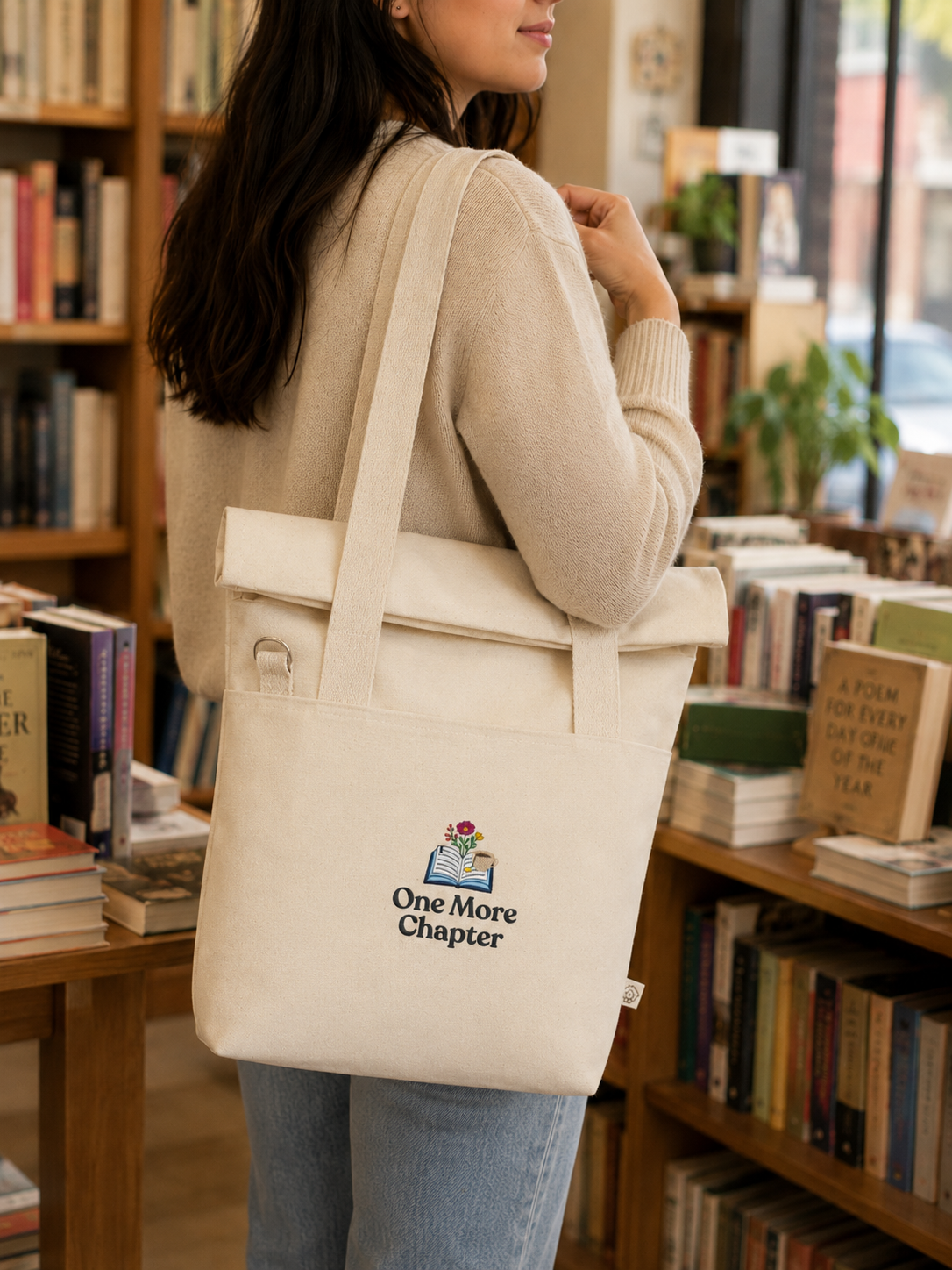Person holding a beige tote bag with 'One More Chapter' logo in a bookstore.