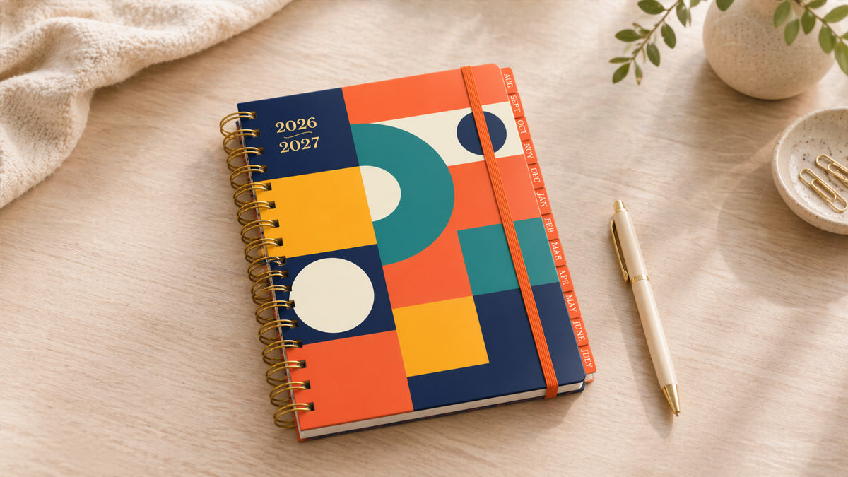 2026-2027 gold spiral colorful planner with geometric design on a wooden surface with a pen and plant.