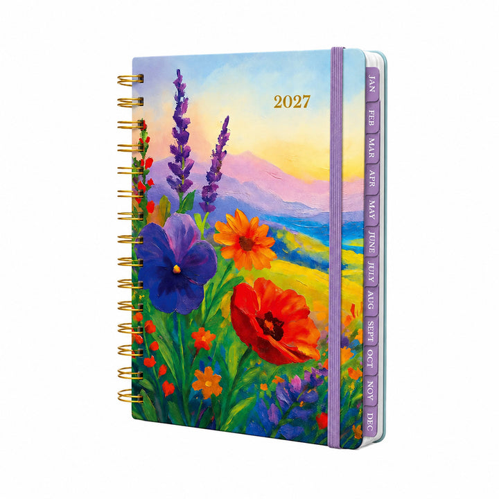 A 2027 spiral-bound planner with a colorful floral landscape cover, featuring purple and orange flowers, mountains, and a sunset sky. The planner has gold-tone spiral binding and purple monthly tabs visible on the side. 