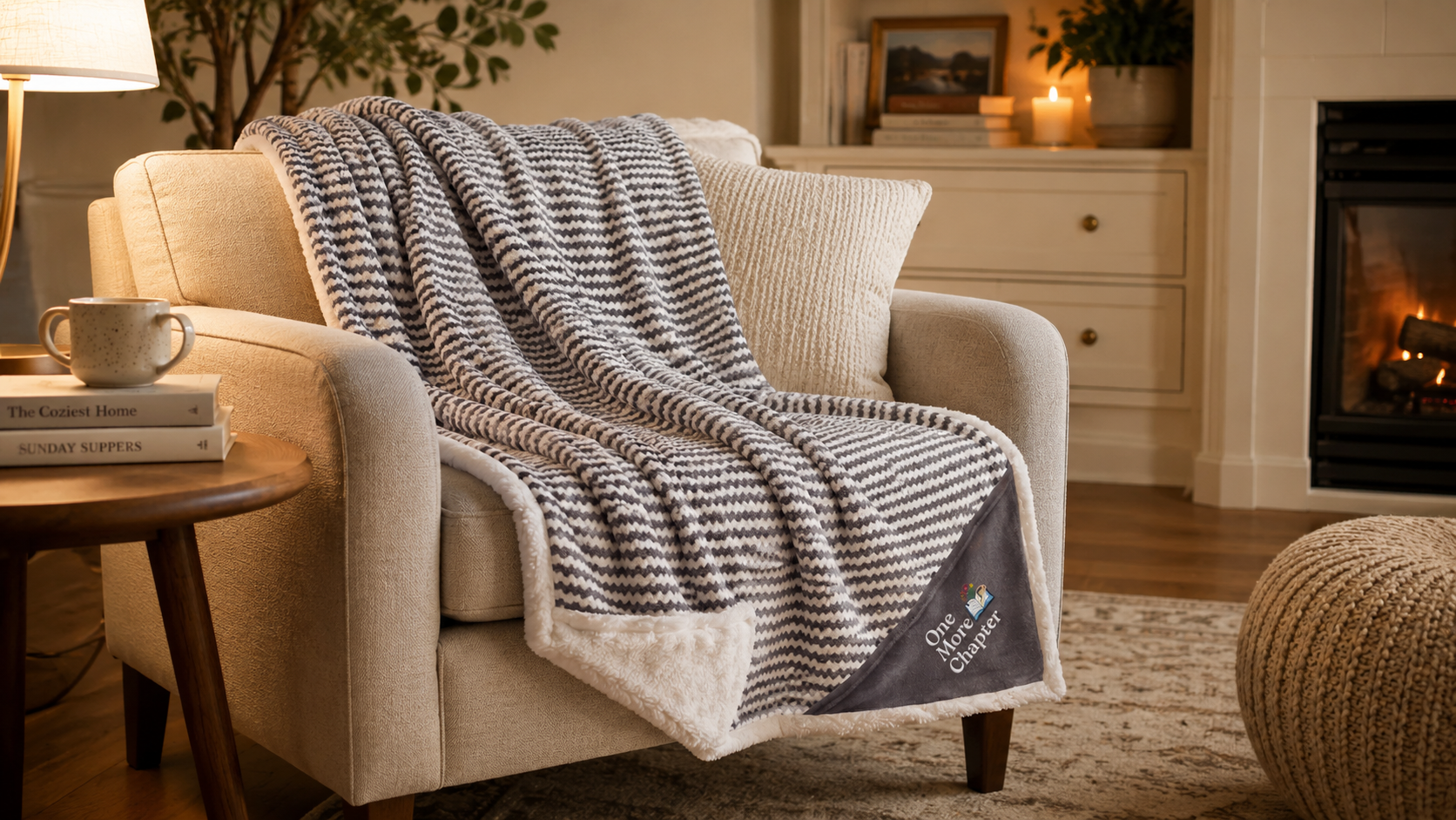 Cozy living room with a beige armchair draped with a textured gray blanket, a small table with a cup and books, and a fireplace in the background.
